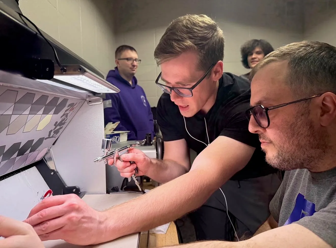 One club member uses an airbrush while another guides a model part inside a spray booth
