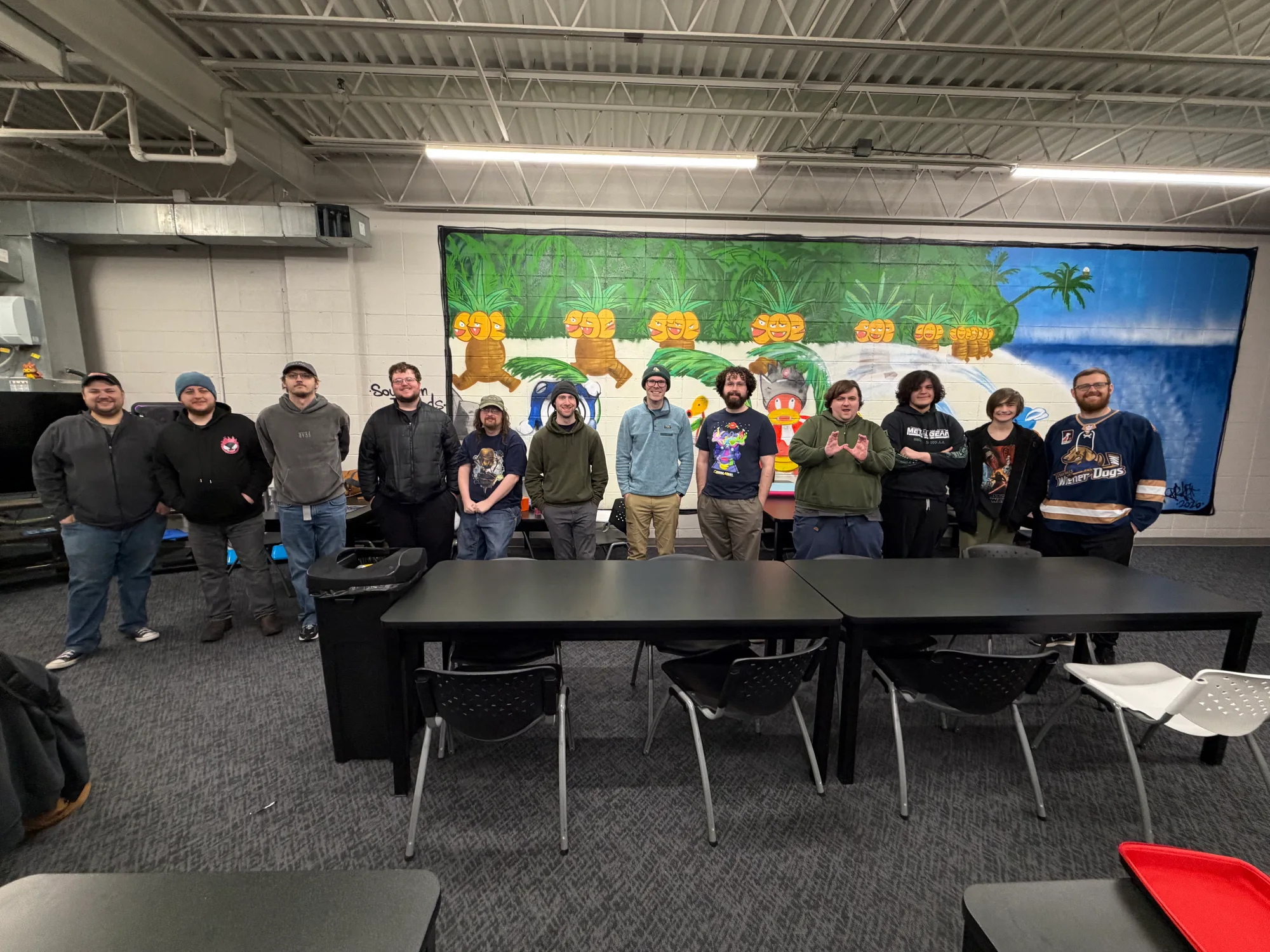 Gundam Club members pose together behind game tables in front of a colorful mural
